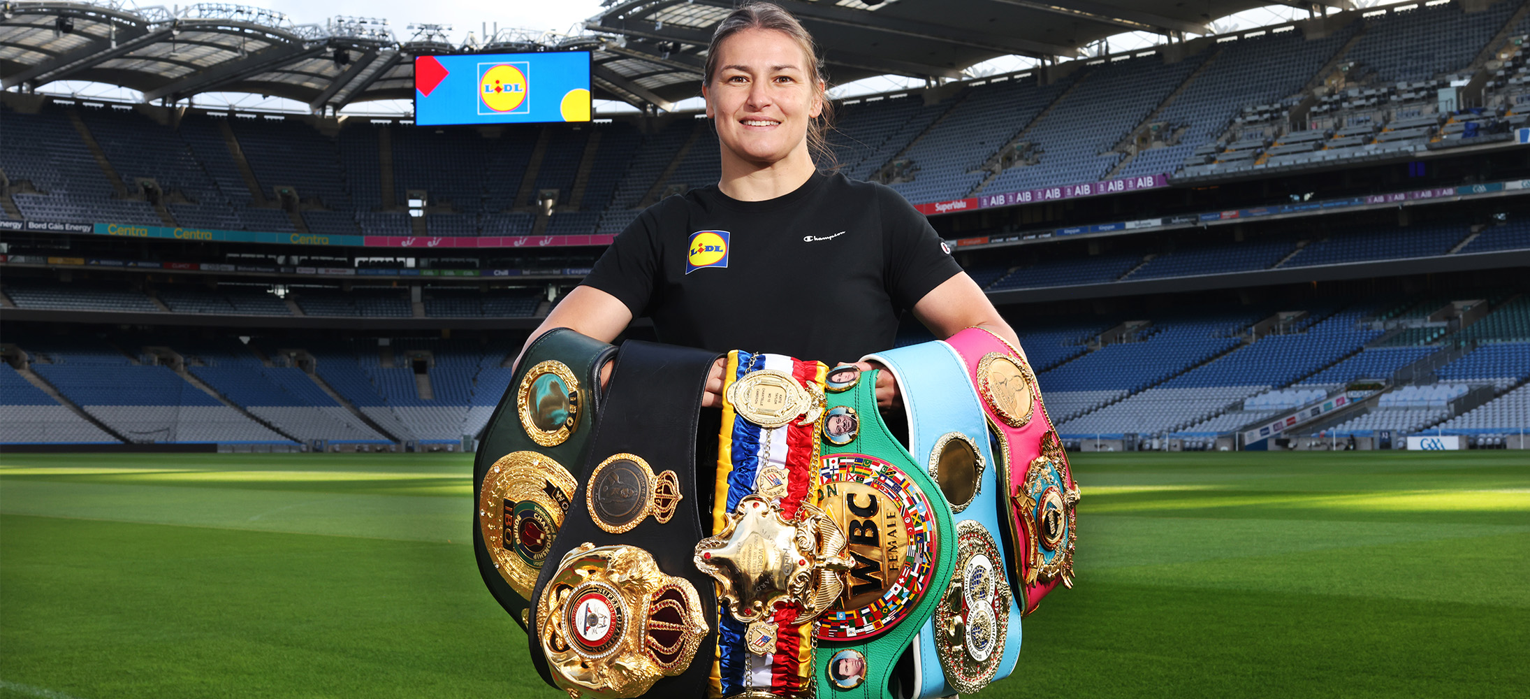 Female boxer holding championship belts in a stadium, with a large screen in the background.